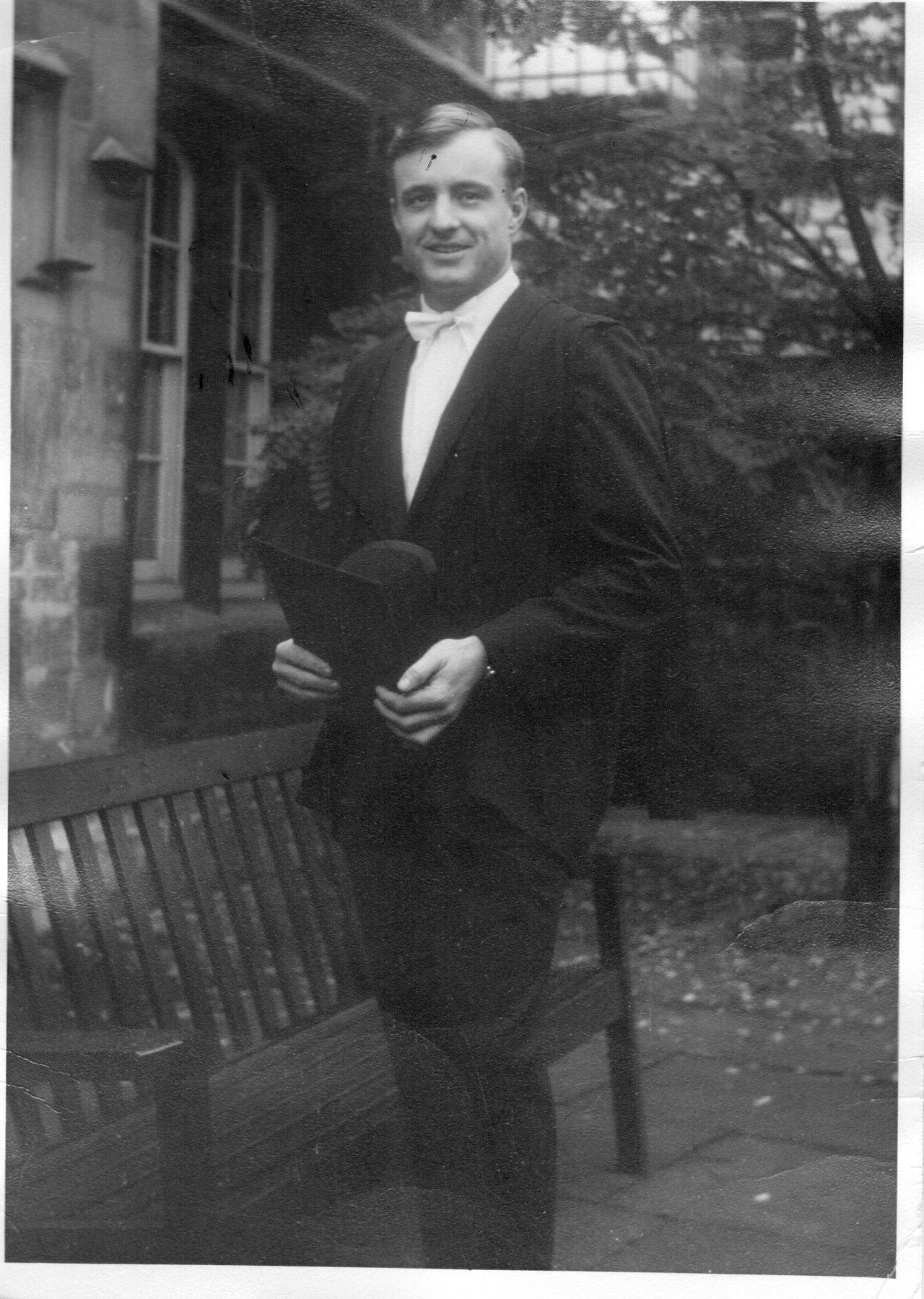 This black-and-white picture shows Richard in a black suit with a sub-fusc, holding a graduation cap in his hands. There's a bench next to him and trees in the background.