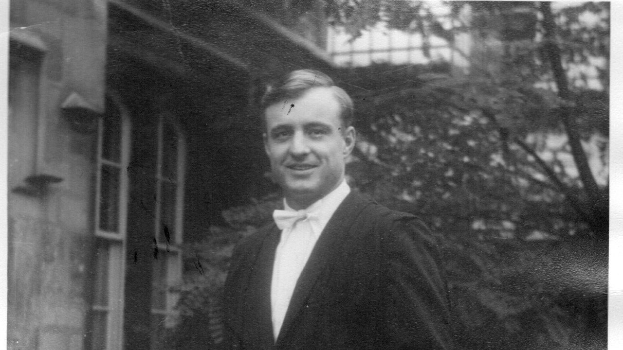 This black-and-white picture shows Richard in a black suit with a sub-fusc, holding a graduation cap in his hands. There's a bench next to him and trees in the background.