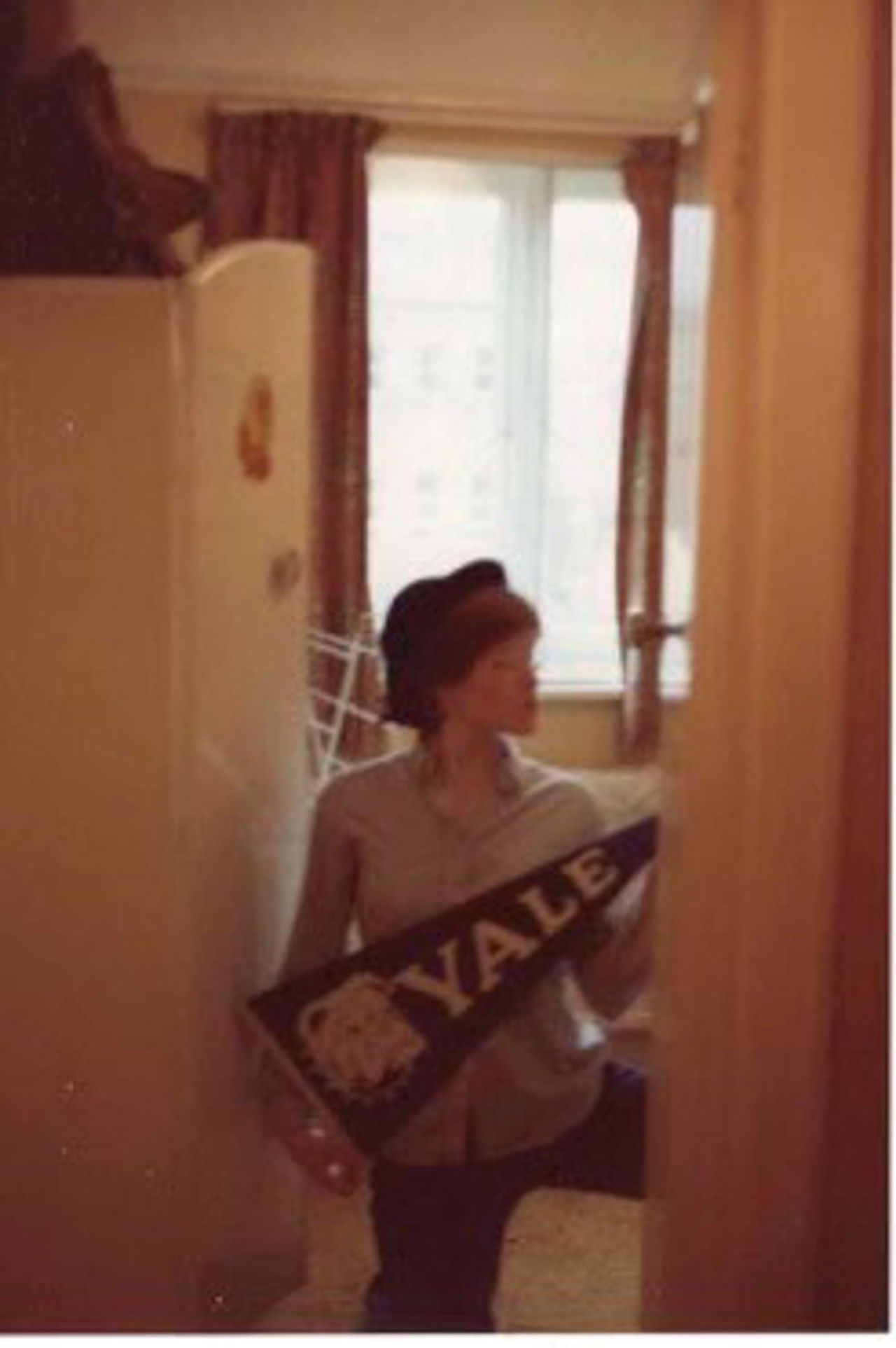 Mary in a shirt and trousers holding a Yale flag in a small room