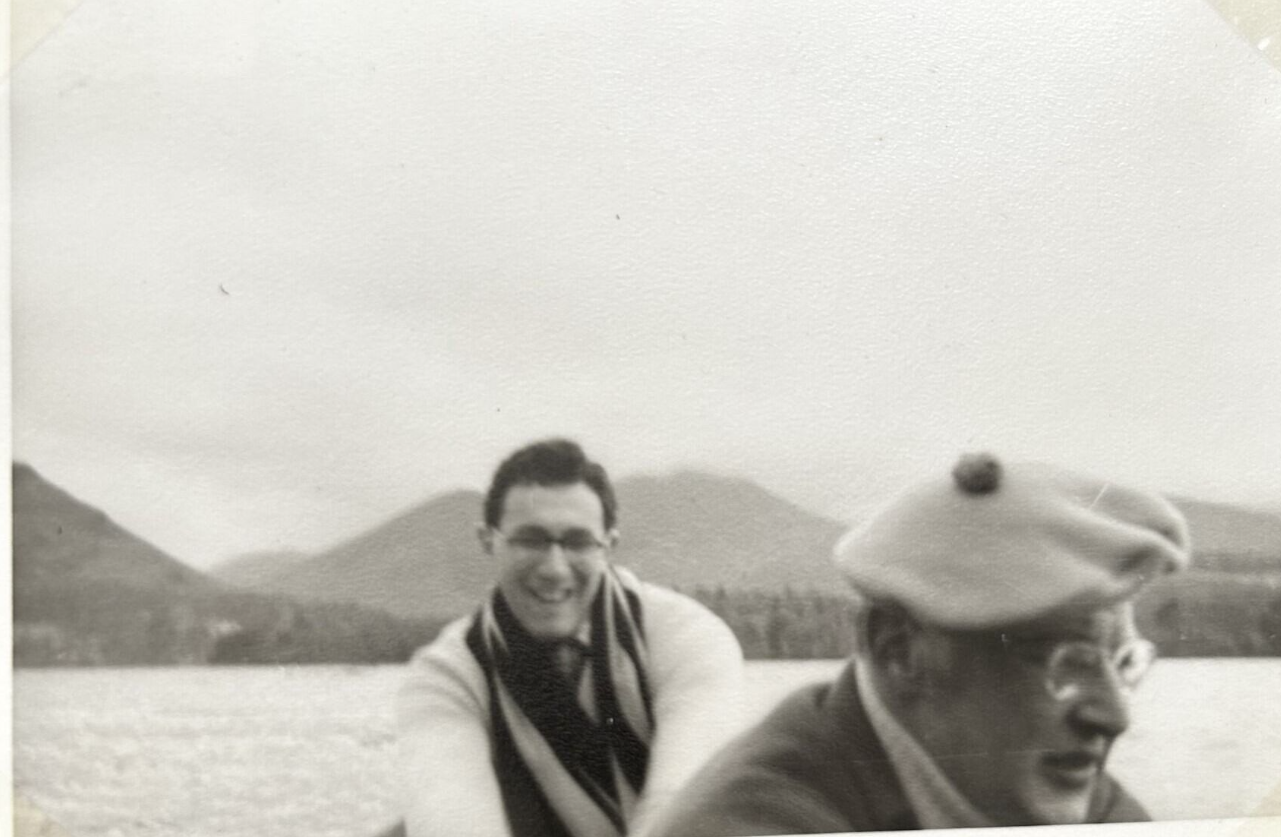 Black and white photo. Two gentlemen appear to be in a boat (unpictured). One sat before Ken wearing a beret, and Ken sat behind him in a scarf and smiling. Behind them there are mountains