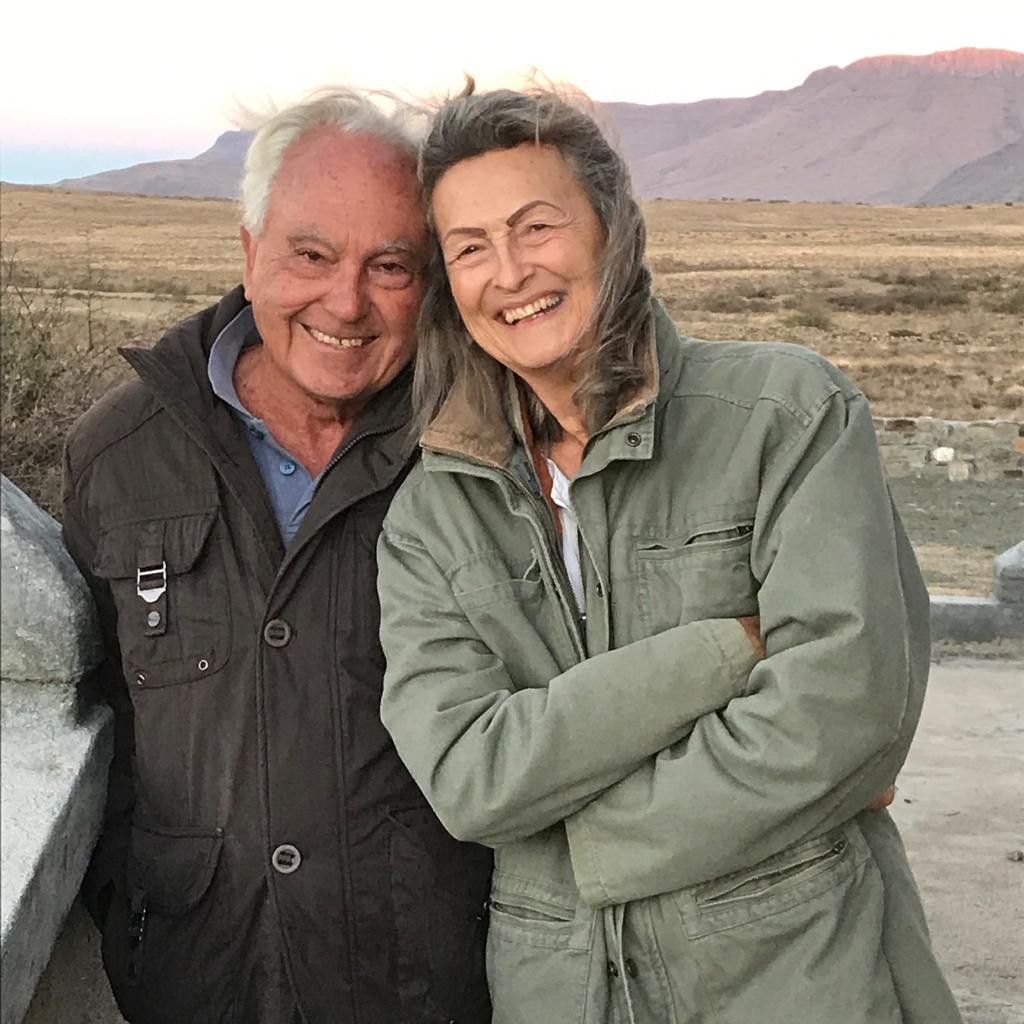 John stood to the left of his wife. They are both smiling widely, and are stood on a form with a field and mountains in the background.