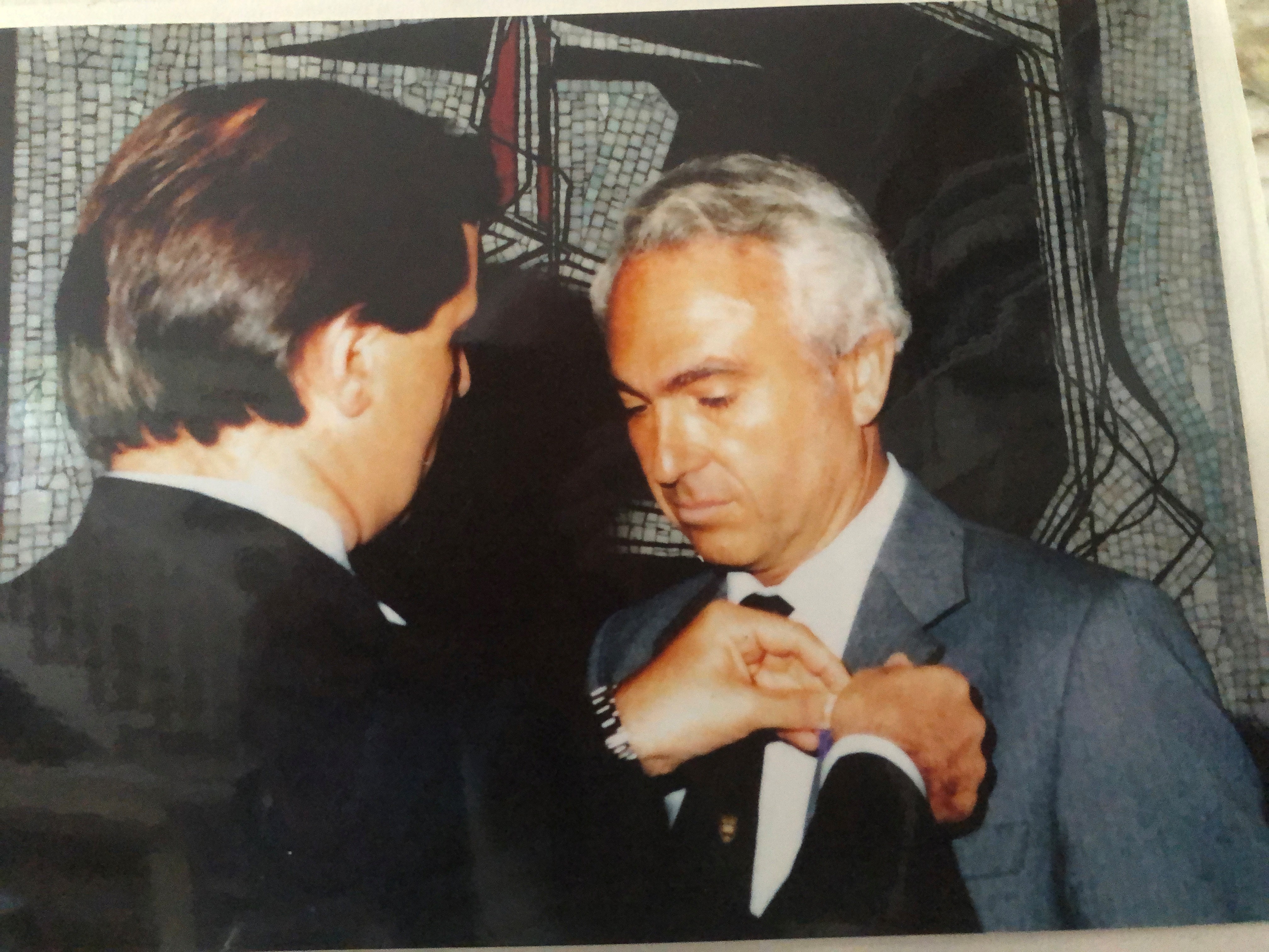 Close-up image of John at the French Embassy. A man with his back turned to us is putting a pin on John's gray blazer.