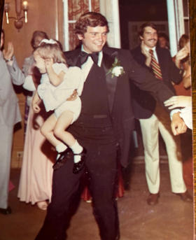 Nick smiling holding a child in his arms, suited, with a crowd behind him.