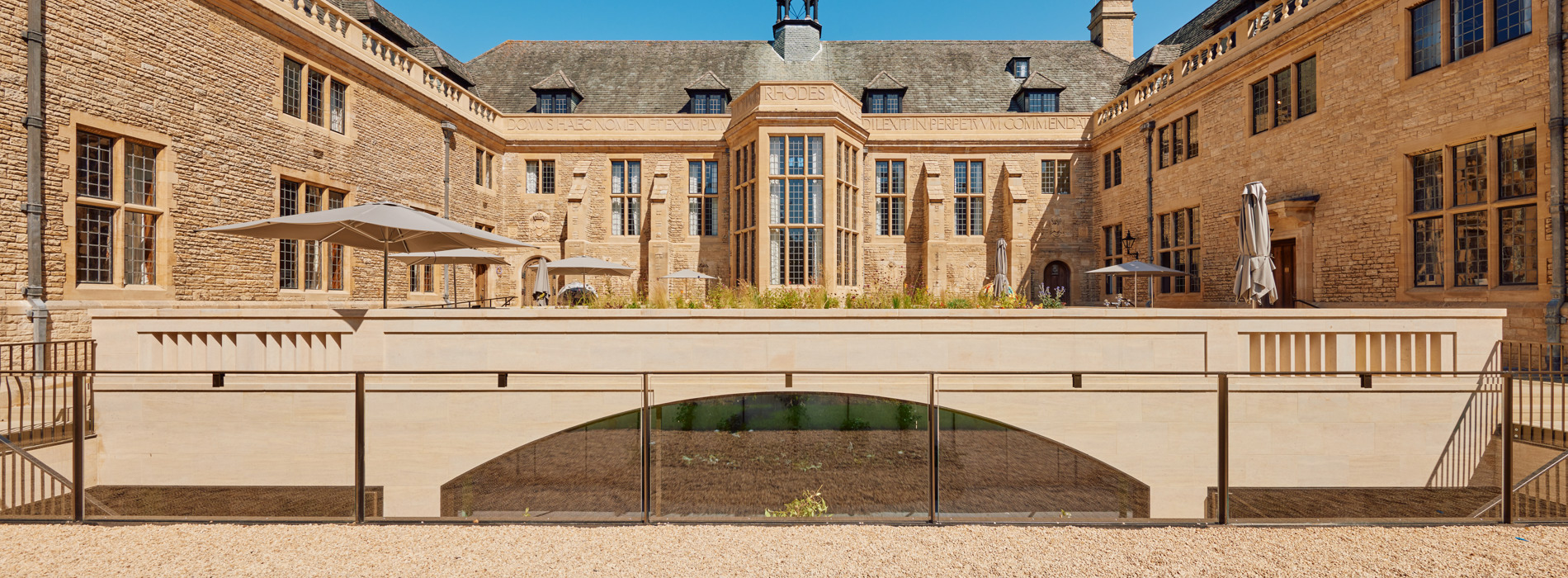 An image of the Rhodes House convening centre exterior. It is outdoors, surrounded by the stone walls of the building, and there are parasols in the seating area. It is sunny and there is a clear blue sky.