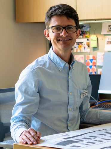 Ben Goodwin sat in a blue shirt at a desk. He is wearing glasses and smiling at the camera.