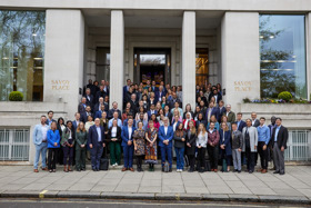 The picture shows a large group of people in formal attire standing in front of a large building whose stone facade is visible behind them.