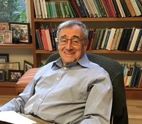 Erich sat in a chair, bookcase behind him. He is wearing a blue/gray shirt and smiling into the camera.