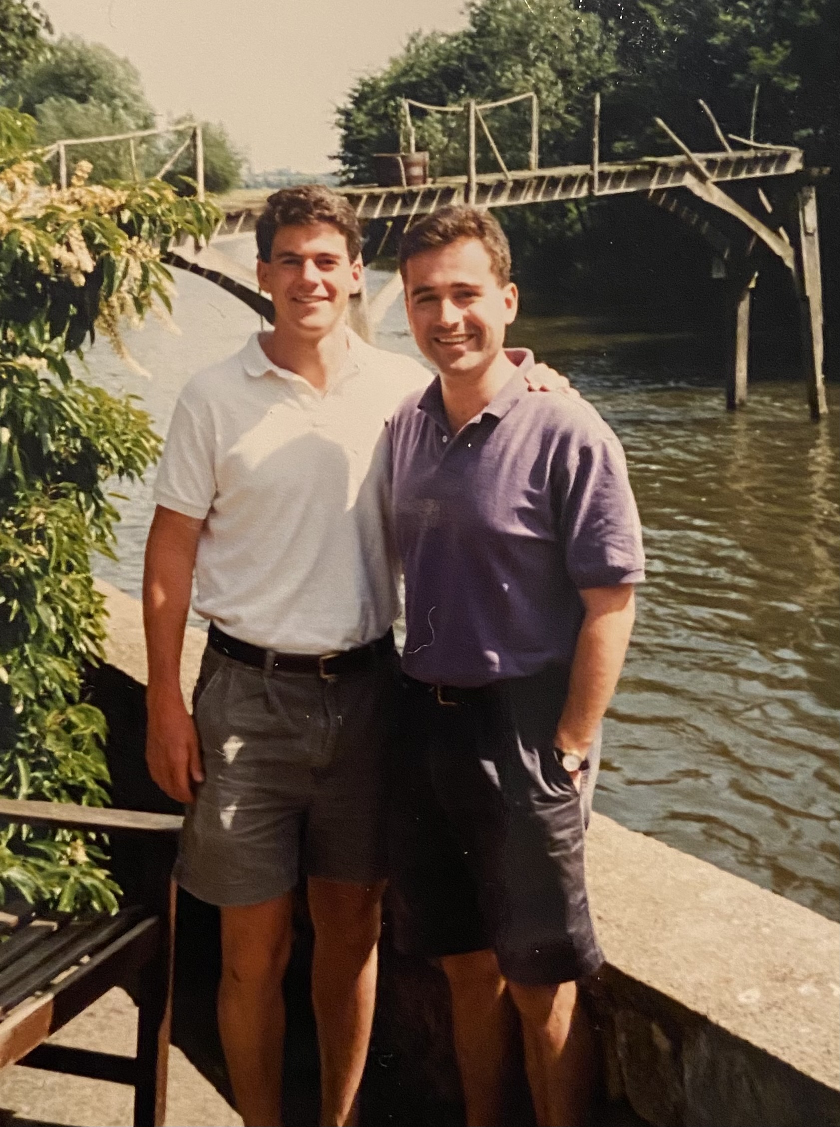 David and Mark are stood smiling with their arms over each other. Both are wearing a polo short and shirts. Greenery is seen around them, and they are stood before a river with a wooden bridge structure.