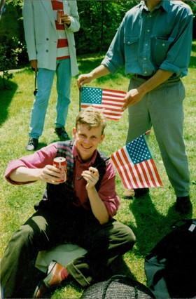 A picture showing a young Mark sitting on a lawn with a can of coke in one hand and an American flag in the other. There are two more men in the picture, one holding an American flag above Mark's head.
