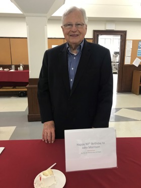 A picture showing John standing in front of a table with a sign that says 'Happy 90th Birthday to John Morrison'