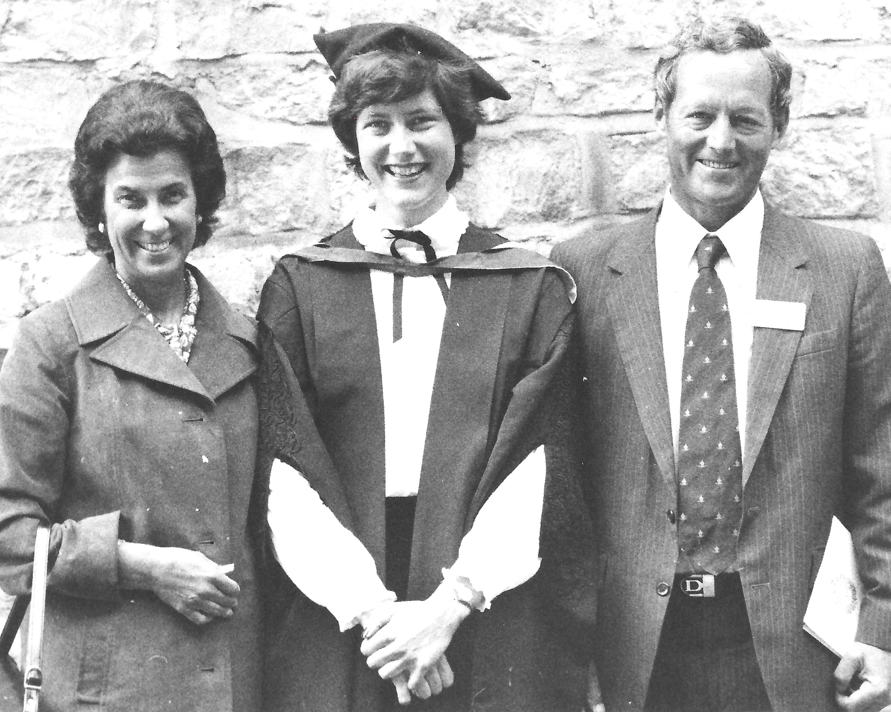 Black and white photo of Margaret stood in the middle of her parents wearing a graduation cap and gown, all smiling into the camera and dressed formally.