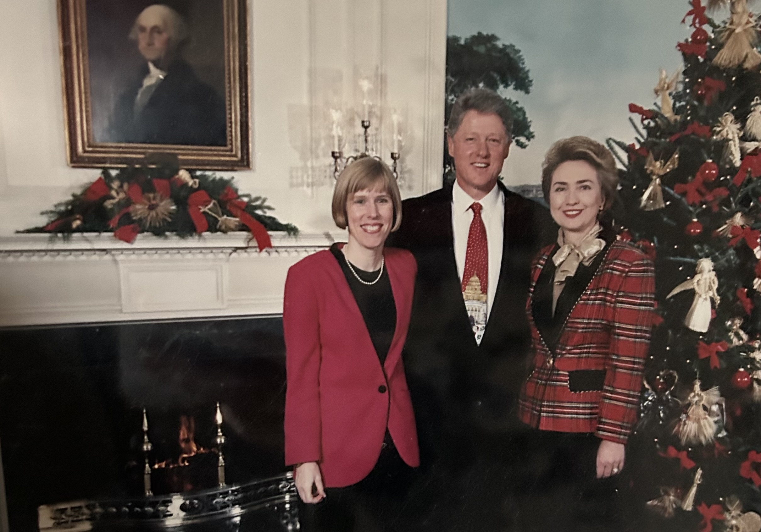 Meredith is pictured to the left of Bill Clinton, and Hilary Clinton to his right. All are wearing red items of clothing, and a Christmas tree and fireplace are seen behind them.