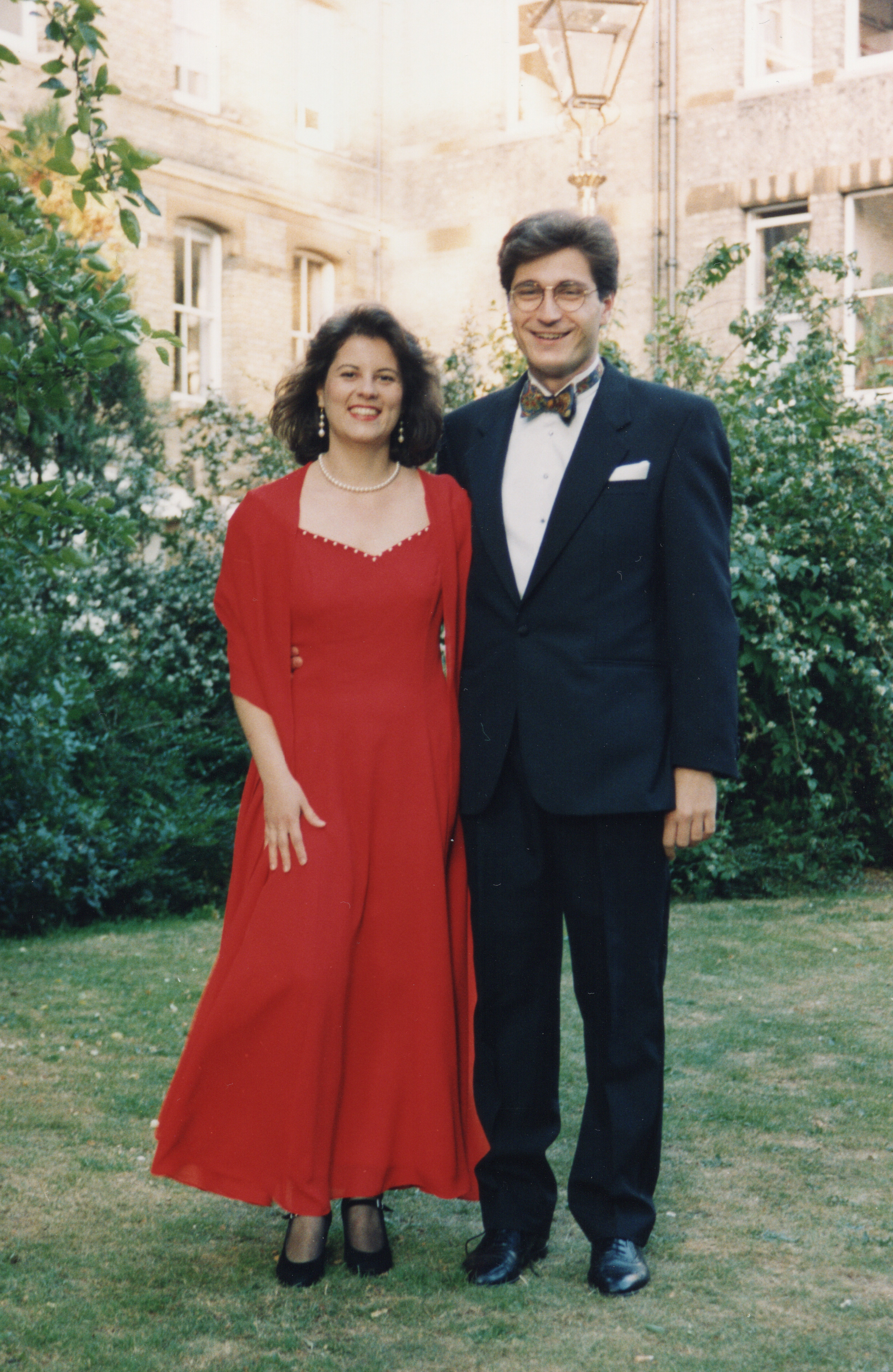 Argyris wearing a suit and bowtie stood to the right of a lady wearing a red gown. They are possing in a college quad.