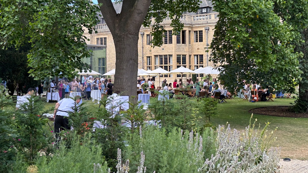 Guests at a Rhodes House BBQ with the chefs preparing food under the trees.