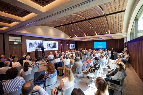 An image of a conference room with dark wooden walls and curved ceilings. It is filled with attendees and a man in a suit is speaking at the front.