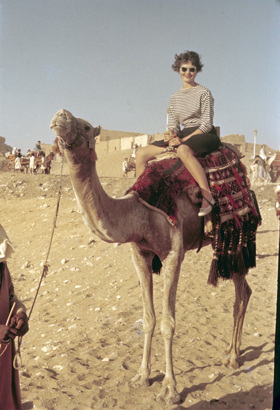 A picture showing Jaynie atop a camel in a desert. She is wearing sunglasses.