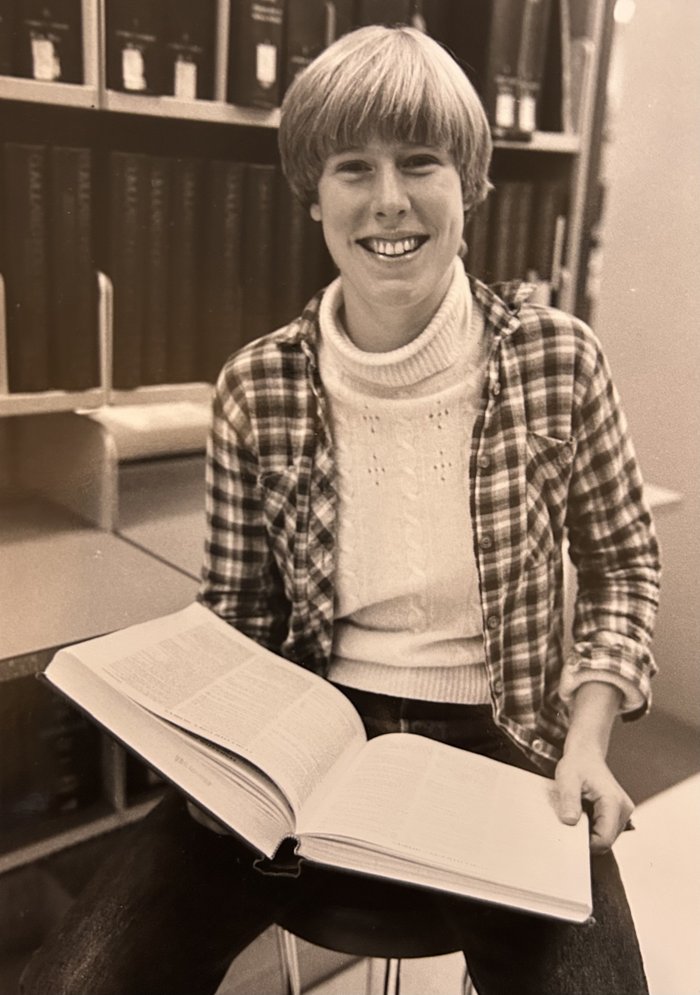 Black and white photograph of Meredith sat on a stool. She smiles, as she holds open a large book.