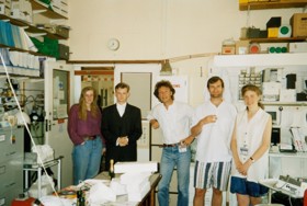 A picture showing Mark in formal attire with his coworkers and supervisor in casual wear. They are standing in a messy office.