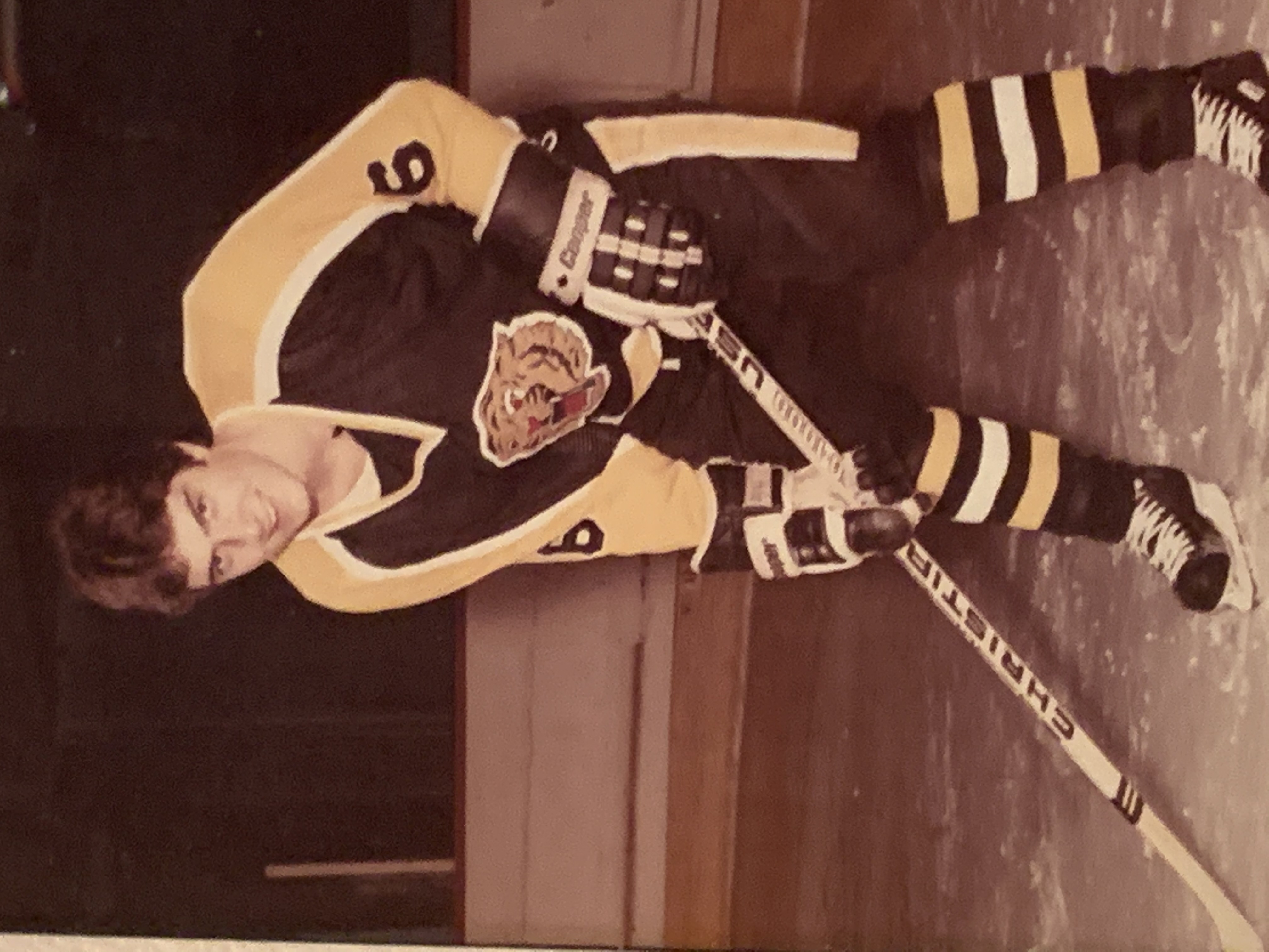 Paul wearing a yellow and navy hockey uniform, he is skating on an ice rink, holding a hocket stick and looking at the camera.