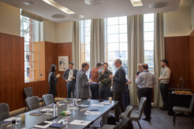 The picture shows Philip standing amongst a group of people. They are all dressed formally. There are large, long windows in the background. There is a table in front of them, and some catering staff are in the room as well.