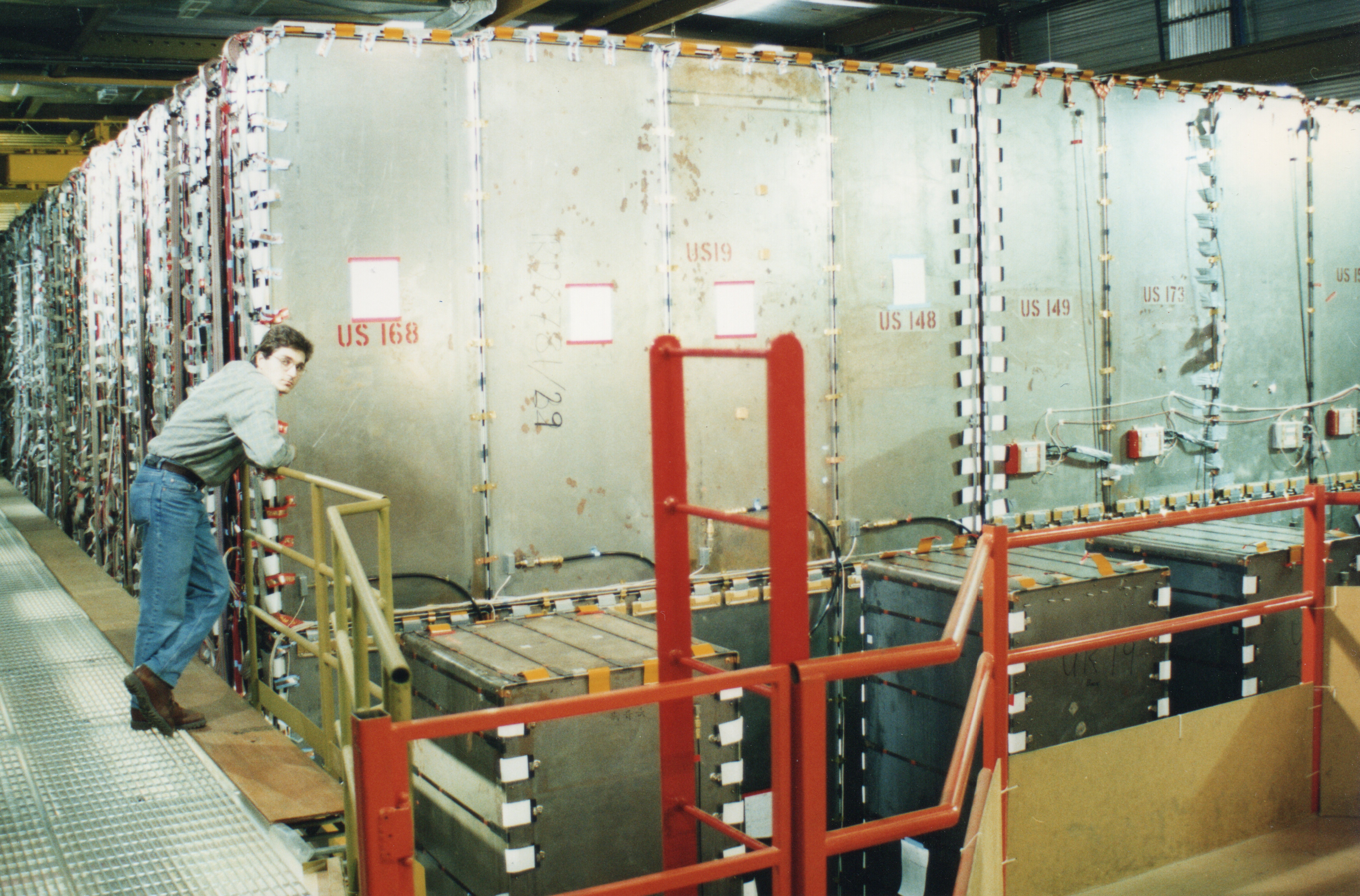 Argyris is stood in a large warehouse with a large metalic structure in the middle. He is leaning over some railing in front of the metal structure which is stamped with the code 'US 168' and other numbers all over.