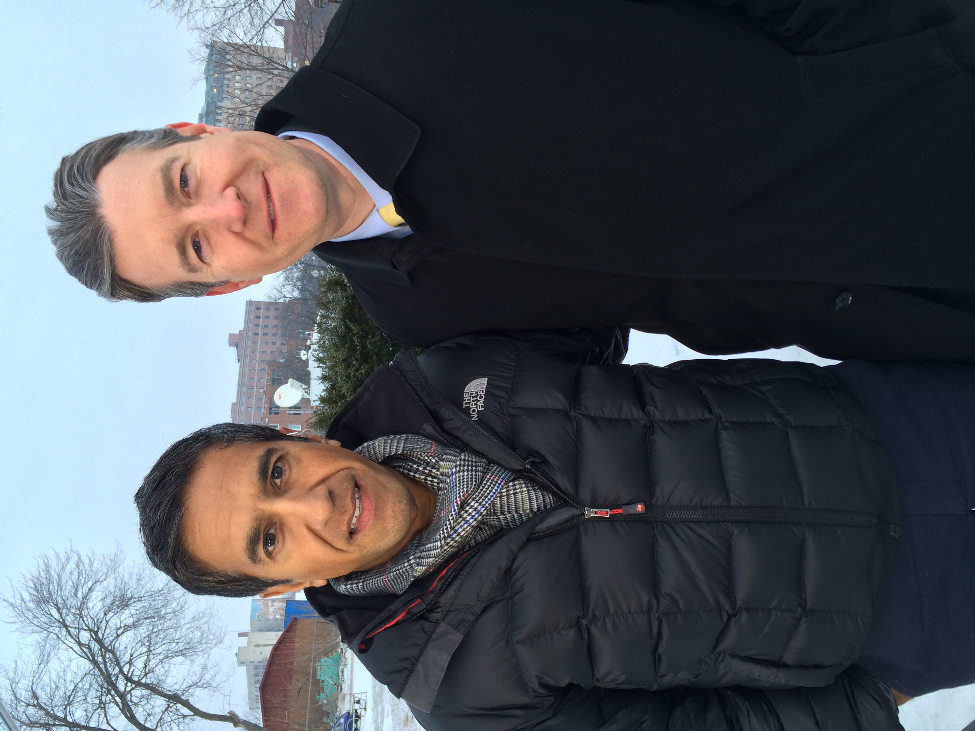 Dayne is stood to the right of Sanjay. Both are wearing black coats, snow and buildings are pictued behind them.