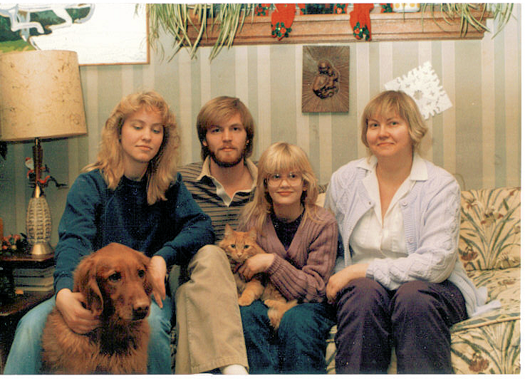 Family of four sat on a floral sofa, green stripy wallpaper behind them. Young woman to the left stroking a dog, Eric as a young man sat next to her, a younger girl holding a cat to the right, and an older woman to the right.
