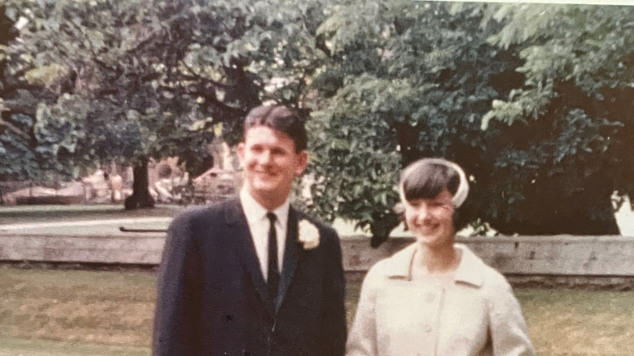 The picture shows Nick in formal attire with his wife in a white dress. They are standing on a lawn.