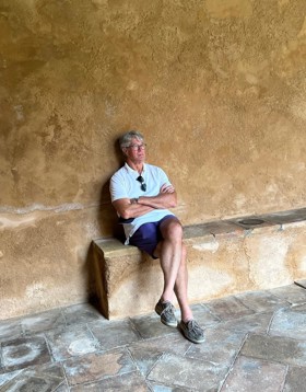 Mike is sat on a concrete bench, there is beige concrete wall behind him. He looks relaxed and is wearing shorts, a white top, and sunglasses clipped onto his top.