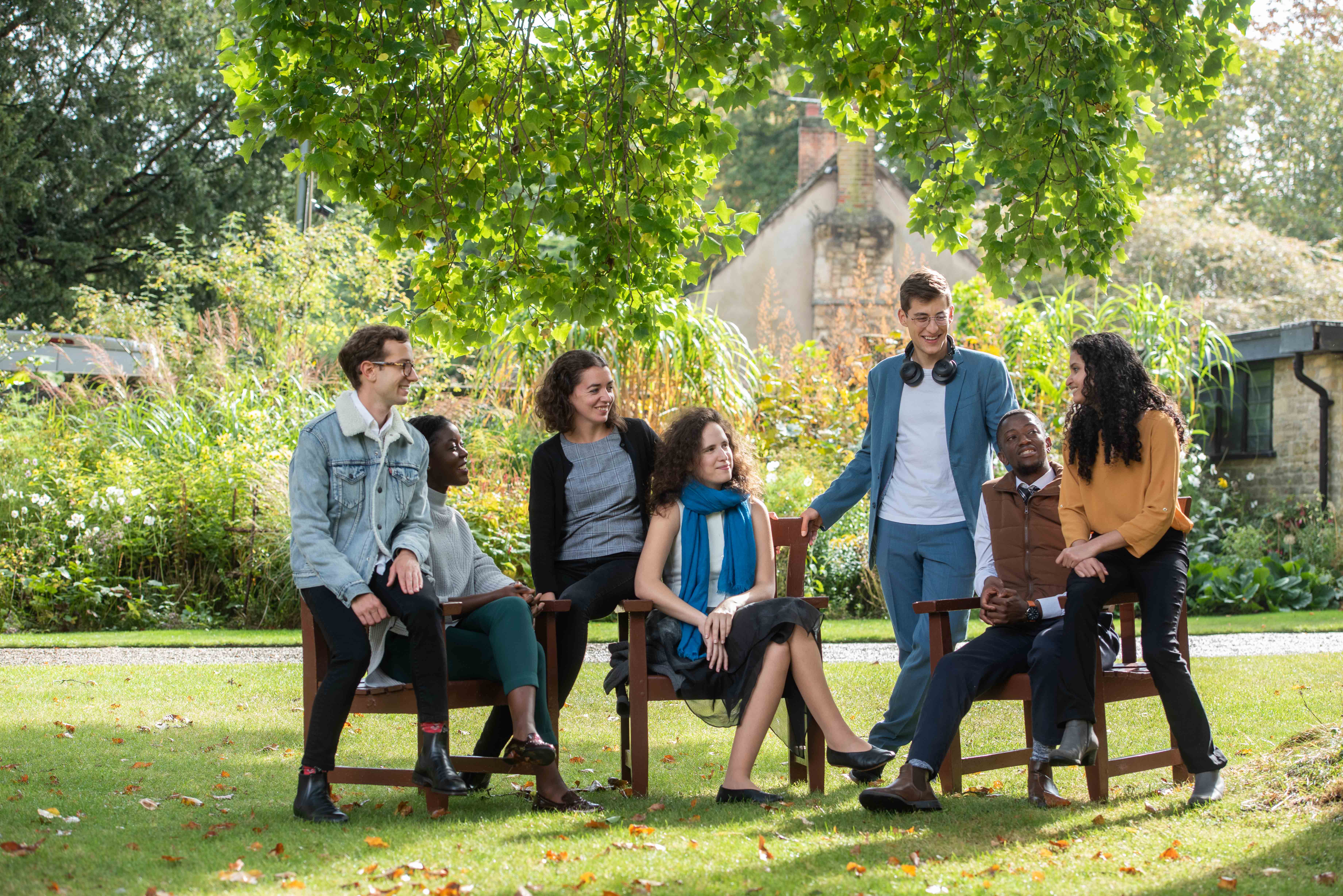 A group of seven Rhodes Scholars alternately sitting on chairs and standing in a row facing the camera. They are outside, on a lawn under a tree in the sun. 