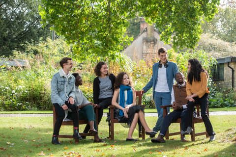 A group of seven Rhodes Scholars alternately sitting on chairs and standing in a row facing the camera. They are outside, on a lawn under a tree in the sun.