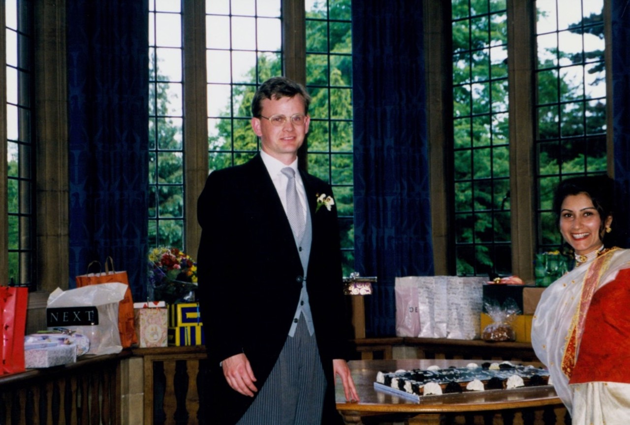 The picture shows Mark in his wedding suit with Shazia in her red-and-white wedding dress with a chocolate cake on the table. There are long windows behind them and gifts on the window sill.