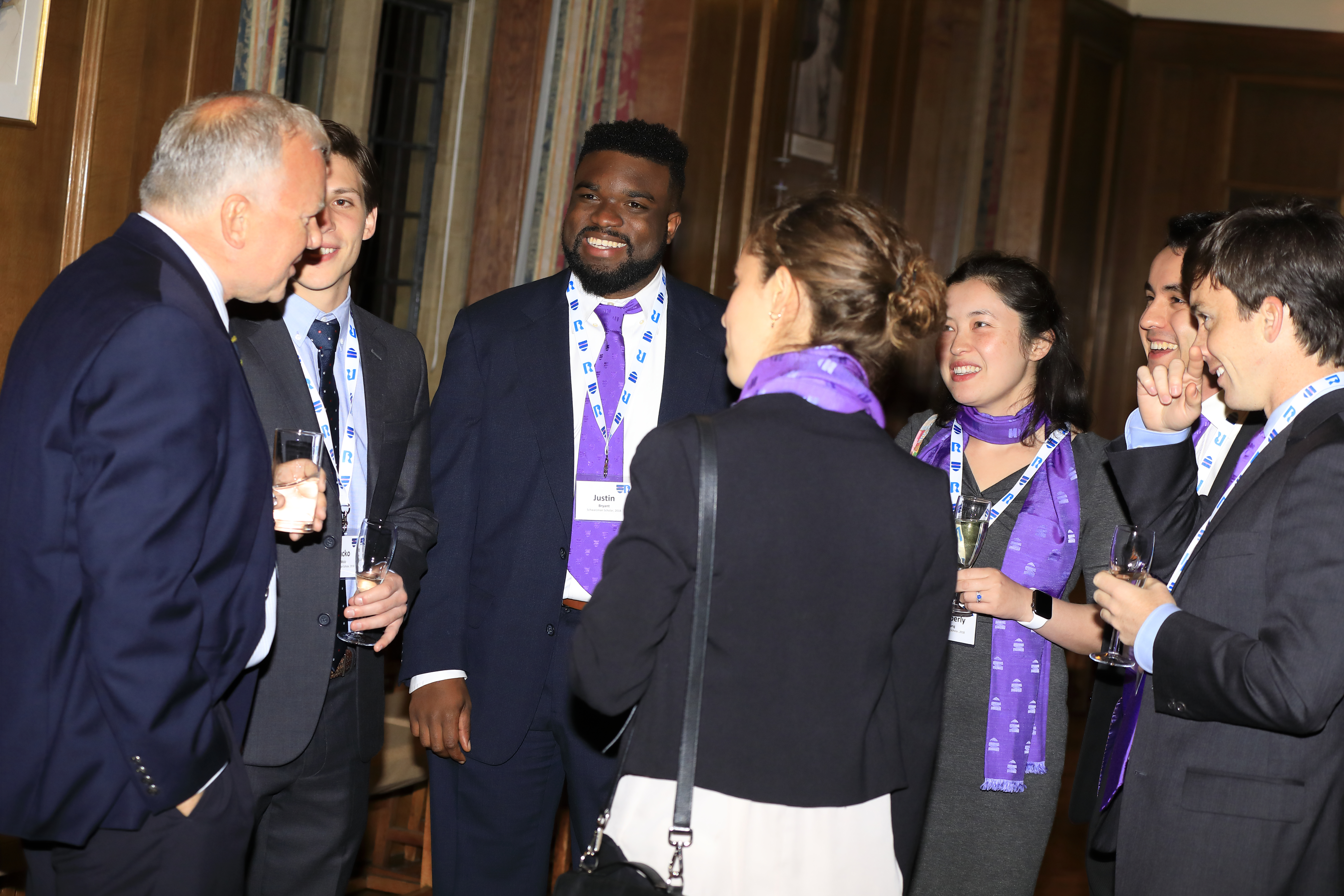 Claus stood to the left talking to a group of Scholars in purple scarves and ties. All are dressed in suits and formal wear.