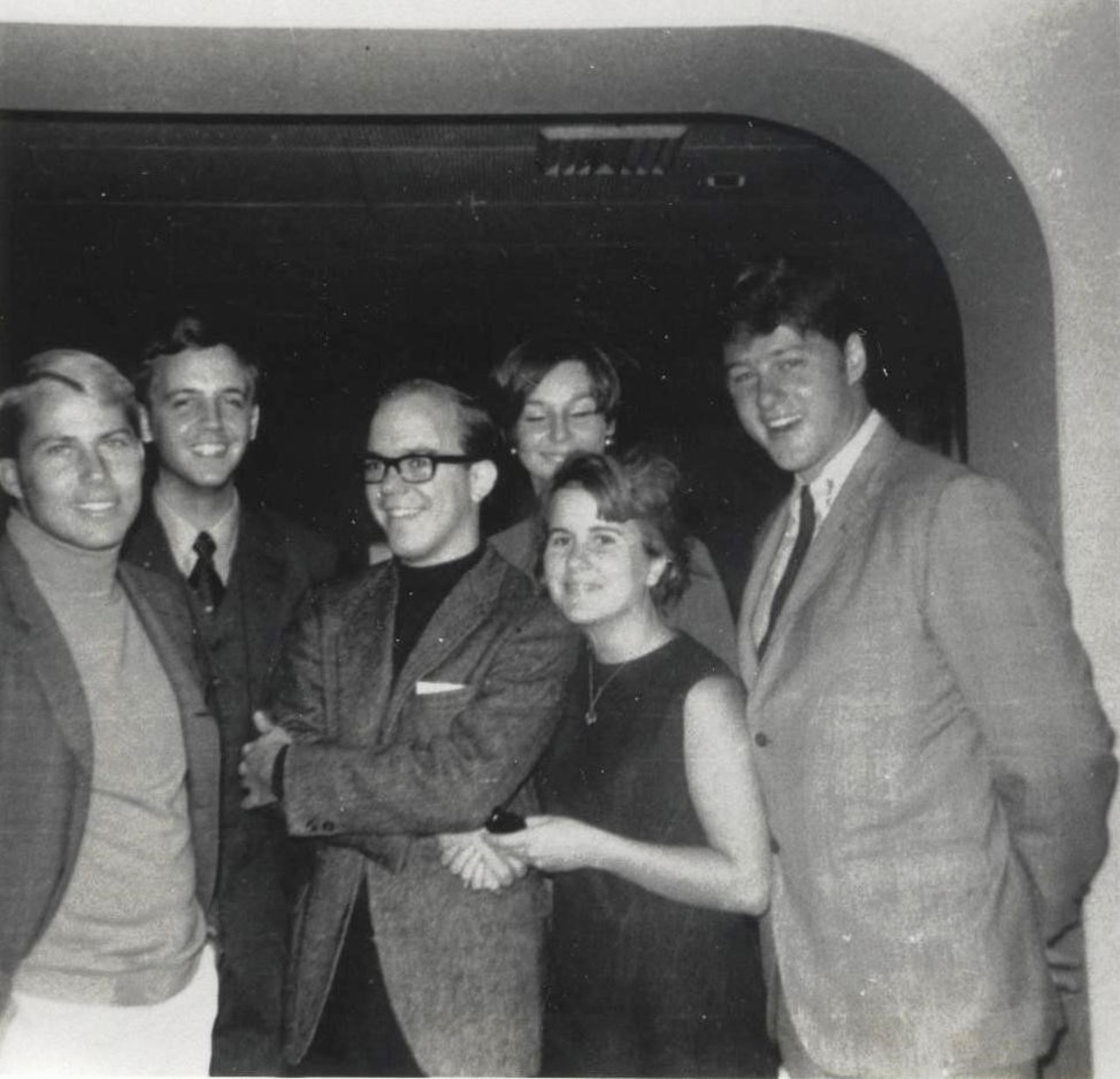 A black-and-white picture showing George with a group of friends inside the USS United States. They are all smartly dressed and smiling. Bill Clinton is standing on the right.