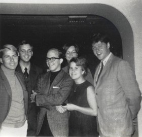 A black-and-white picture showing George with a group of friends inside the USS United States. They are all smartly dressed and smiling. Bill Clinton is standing on the right.