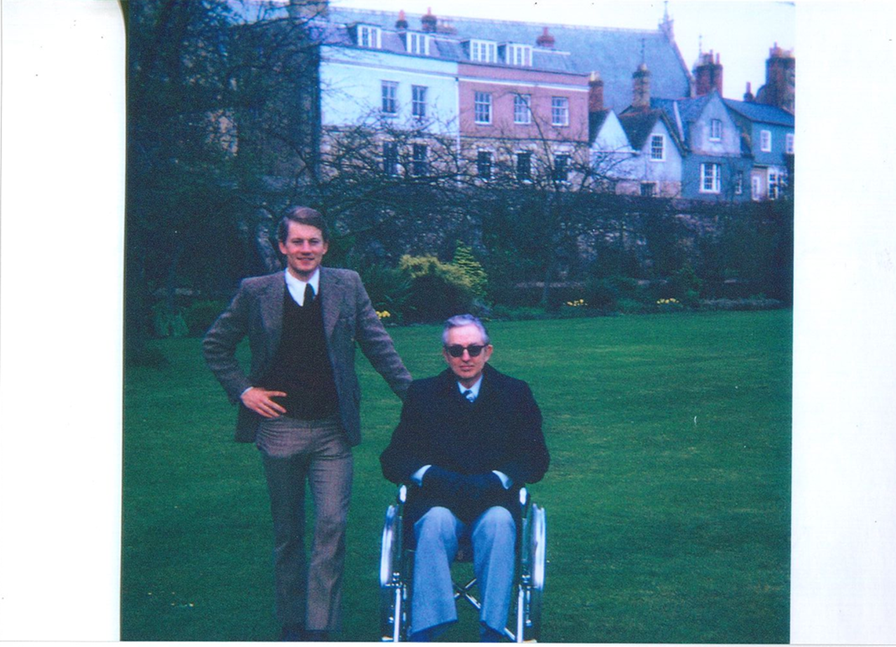 The picture shows Mike in a suit standing next to his dad who is in a wheelchair. Merton buildings are in the background.
