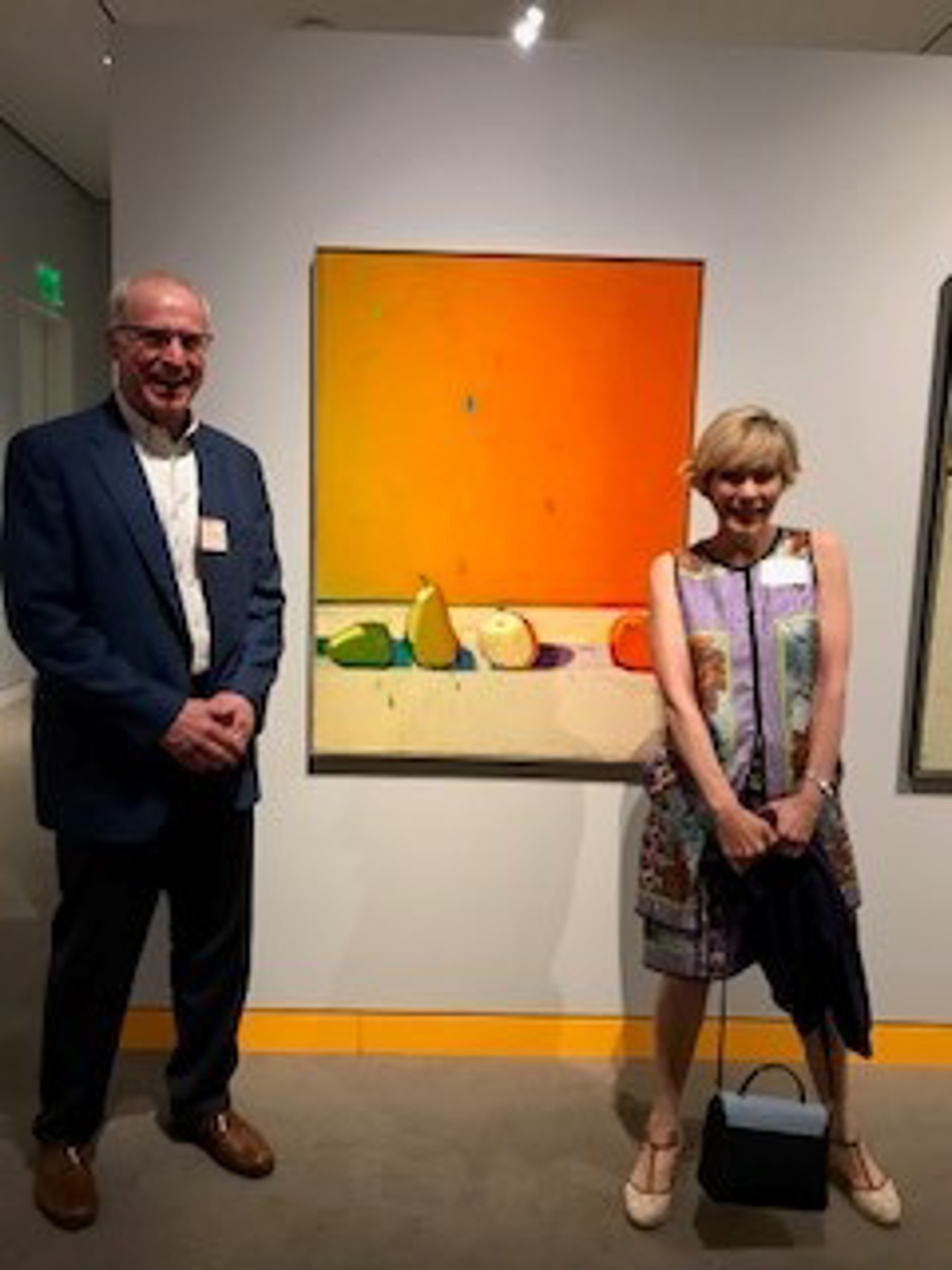 A picture showing Mary next to an orange painting of a few fruits. There's a man in a suit standing next to her.