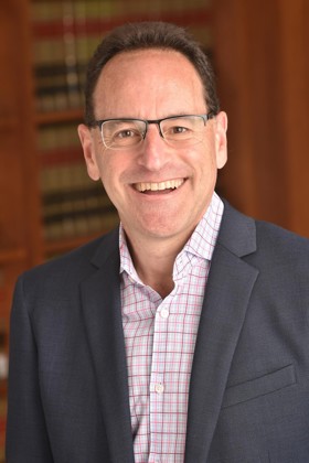 Jonathan in a gray blazer and check shirt, wearing glasses, and smiling into the camera.