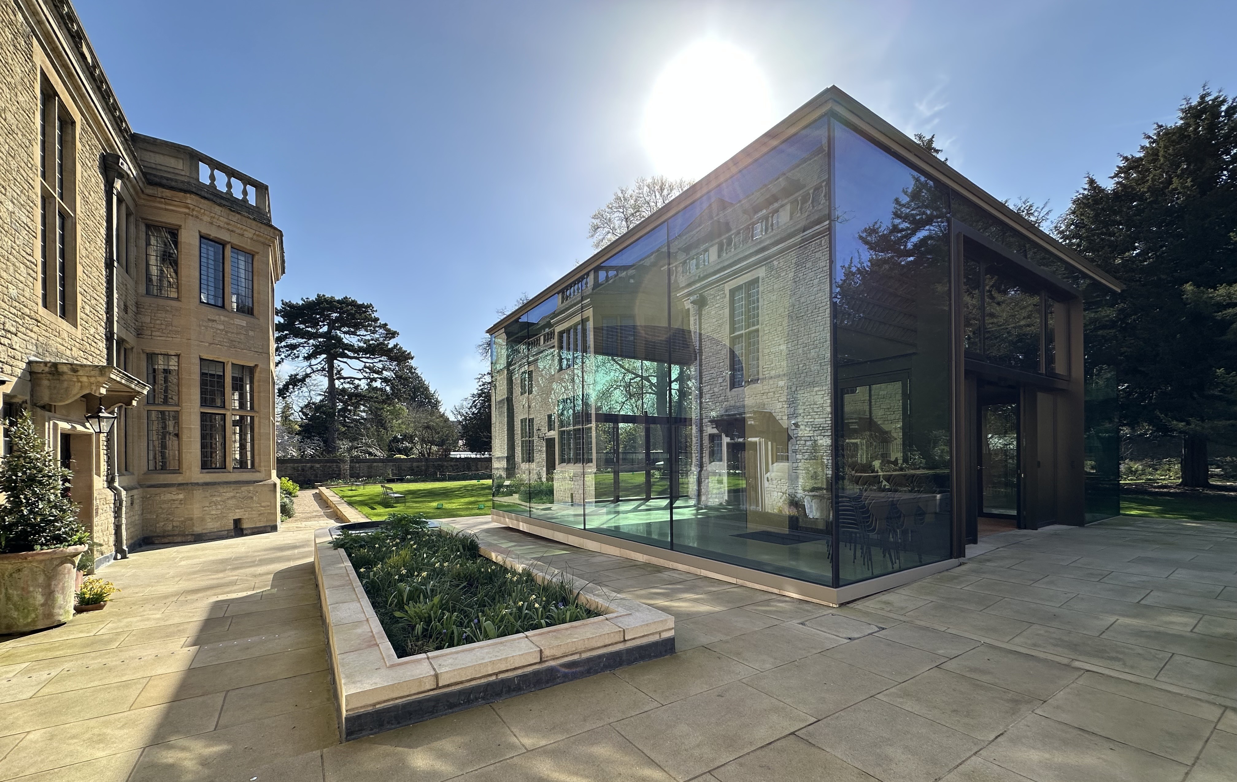 Rhodes House Glass Pavilion with a reflection of Rhodes House on the glass wall and the lawn in the background.