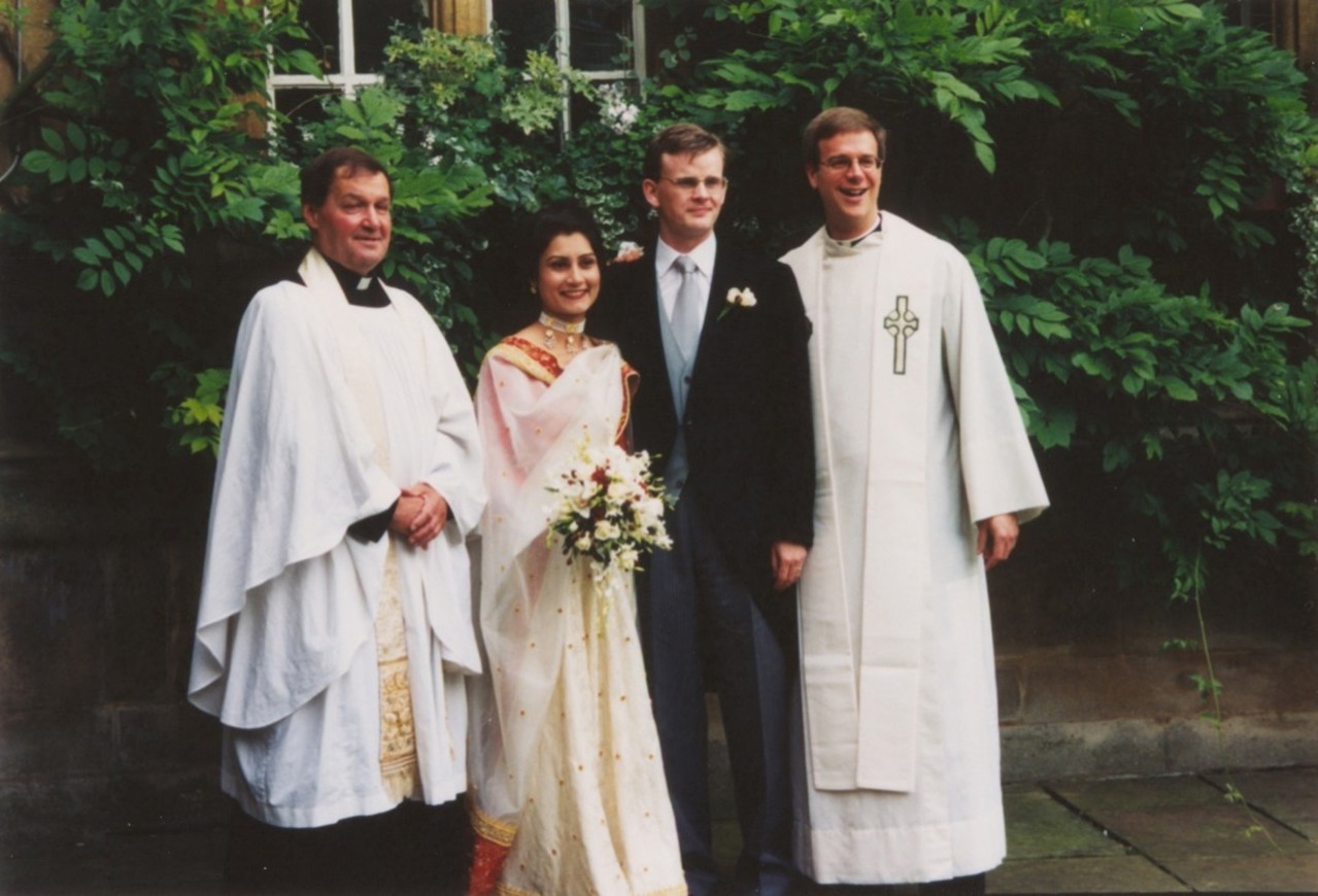 The picture shows Mark, in a suit, and Shazia, in a white gown, with two priests in white robes on either side of them. Thick green foliage in the background.