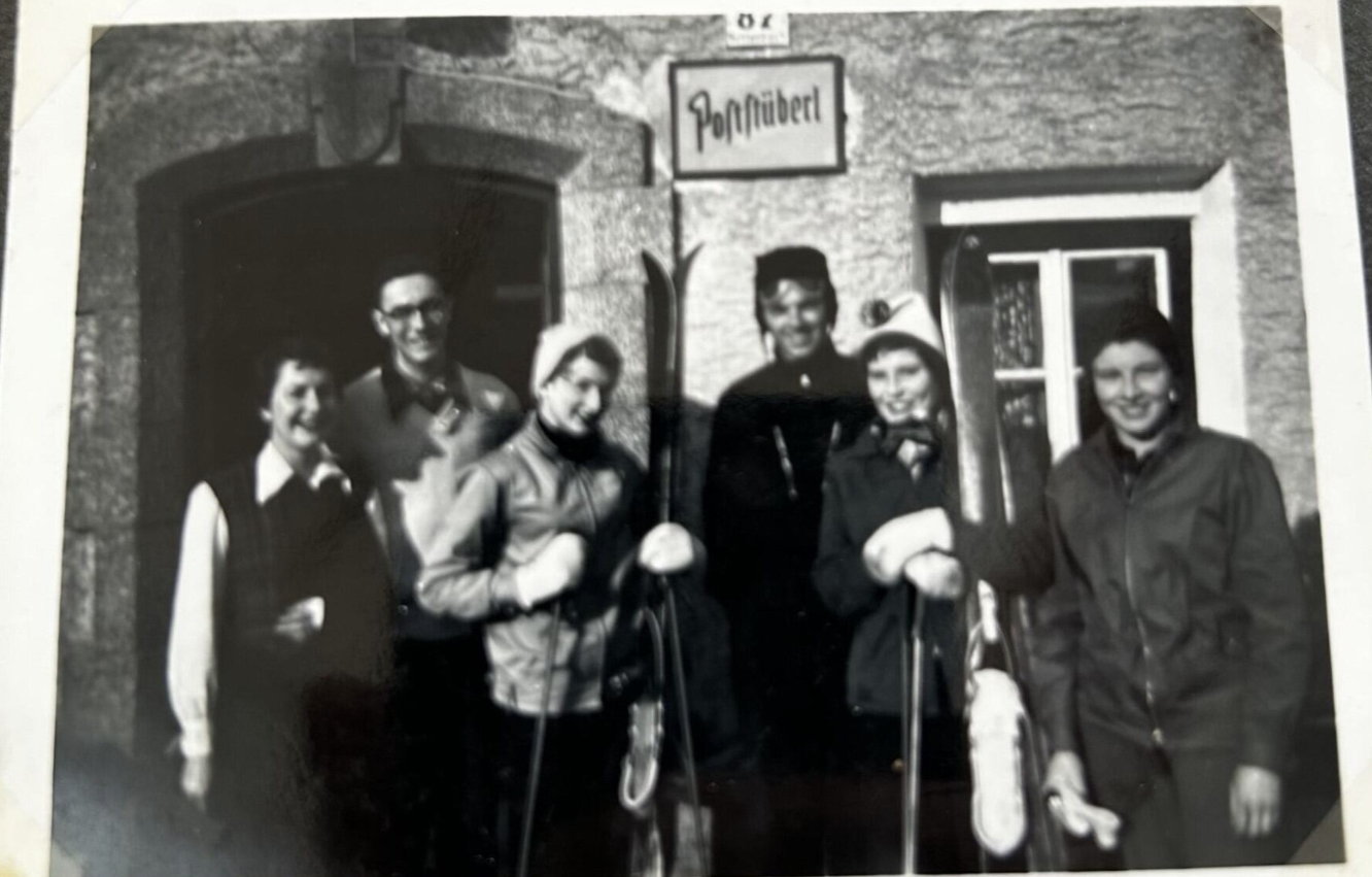 Black and white photograph of a group of young people sdtood before a building. Some are holding ski gear