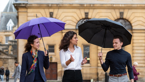 Three students walking outside with the Rhodes House building in the background. Two of them have umbrellas. 