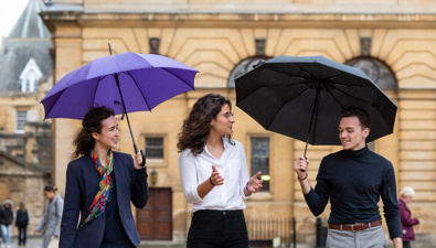Three students walking outside with the Rhodes House building in the background. Two of them have umbrellas.