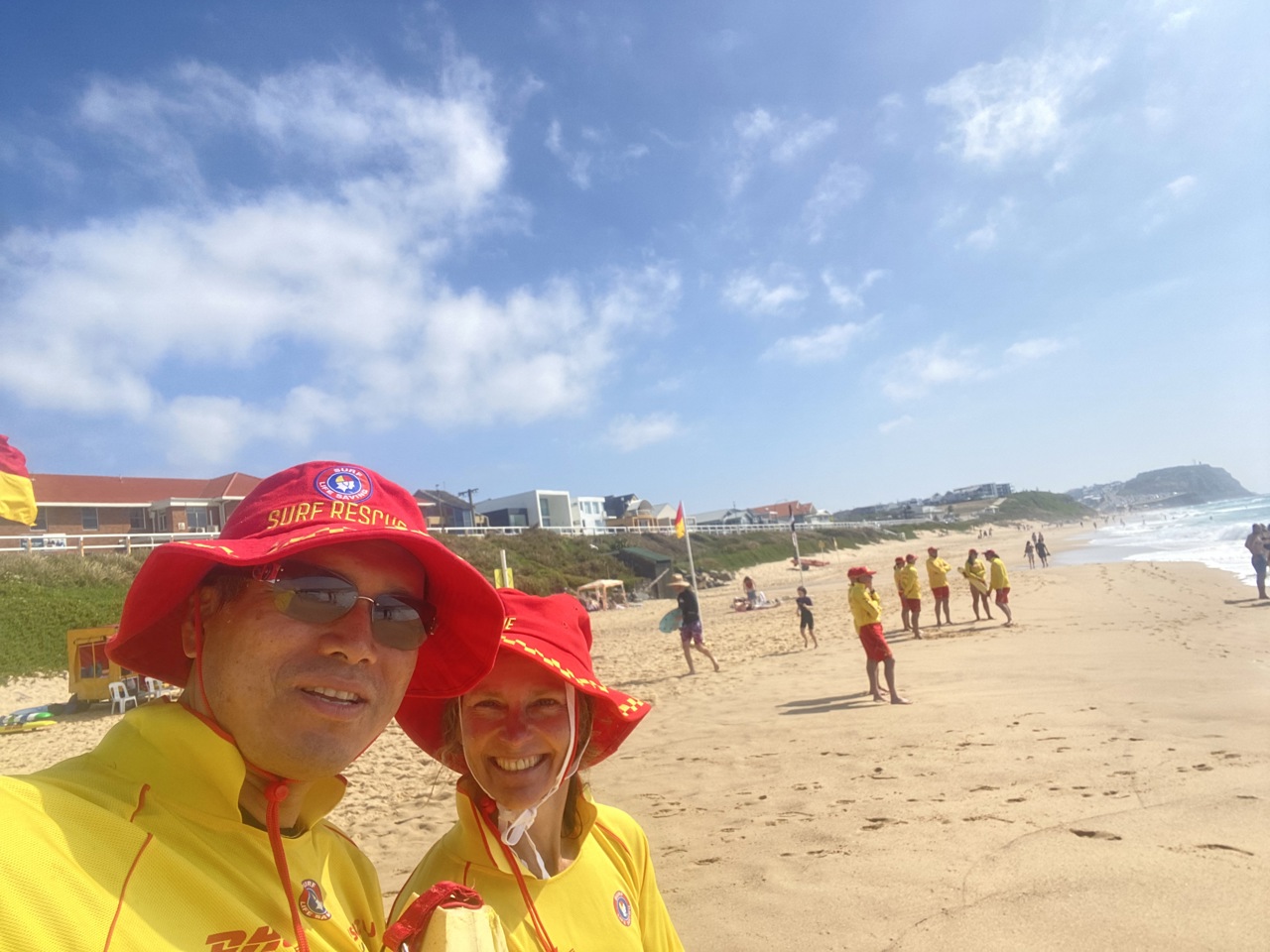Selfie of Jenny and friend on a beach, blue sky and water, both wearing bright yellow and red outfits.
