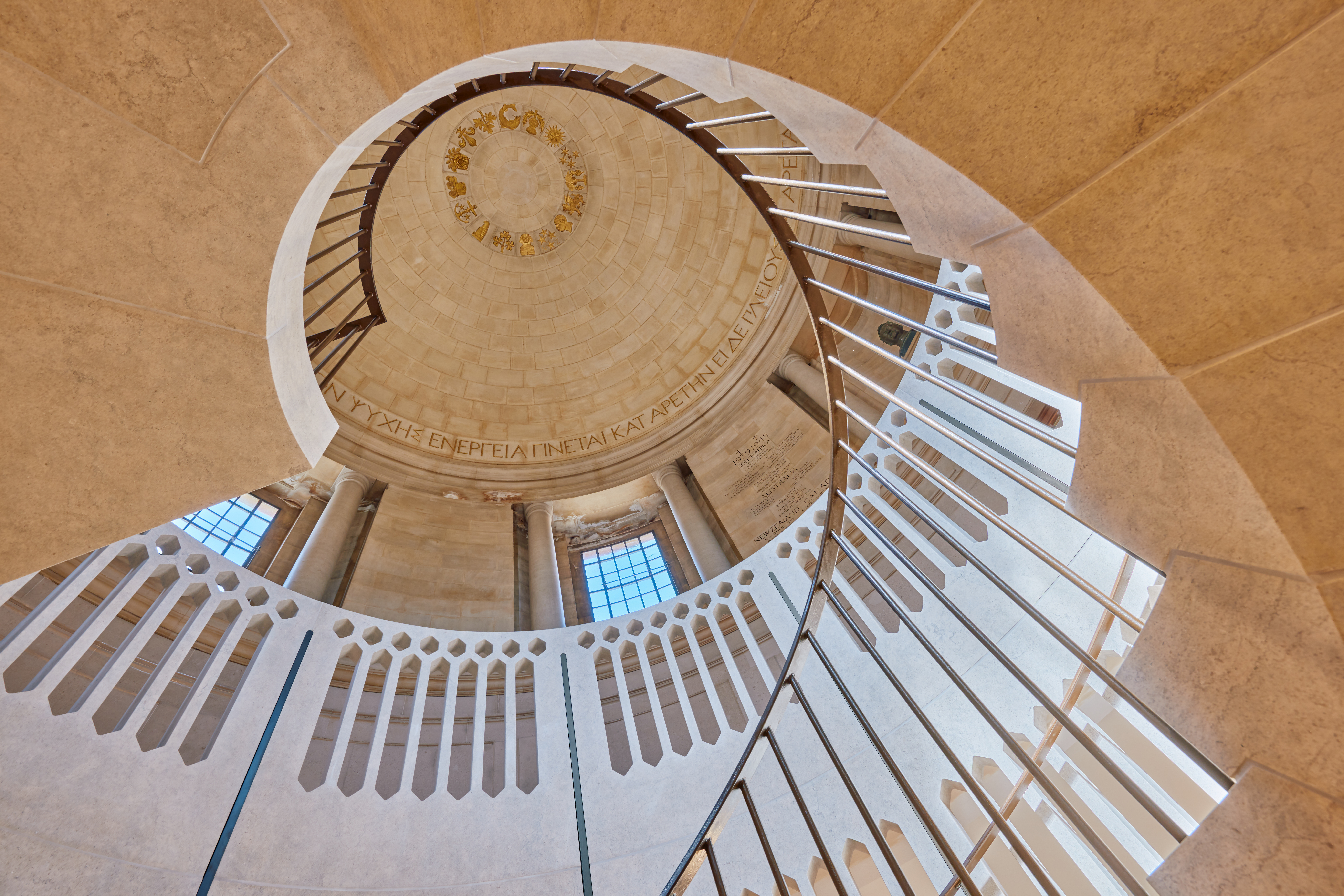 An image looking upwards to the Rhodes Rotunda through a spiral staircase. The ceiling is domed, and the staircase is built of sand-coloured stone. 