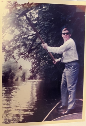 A photo of Bob with a paddle in hand and dressed in smart attire with sunglasses, punting on the river Cherwell