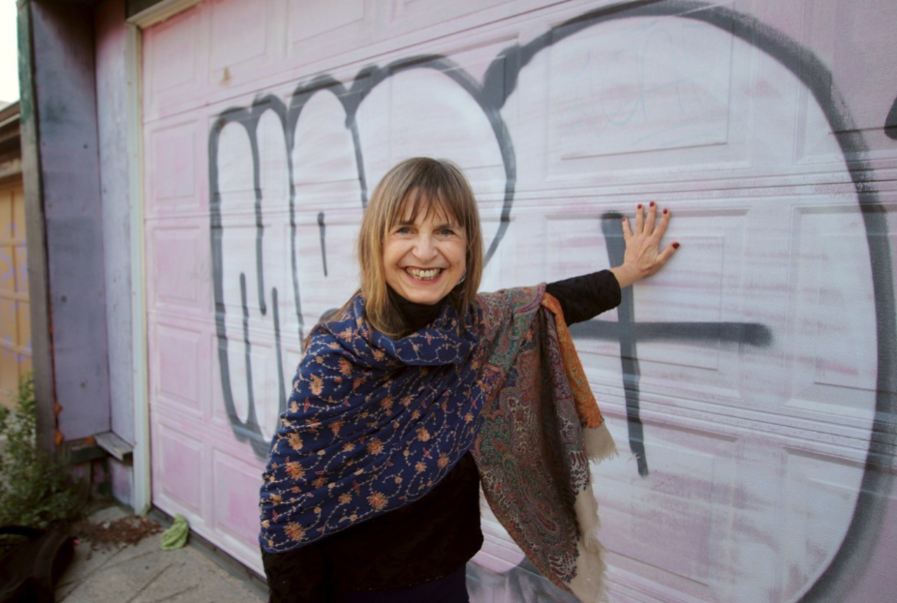 Banuta smiling into the camera, leading against pink and white graffiti on a garage door.