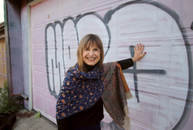 Banuta smiling into the camera, leading against pink and white graffiti on a garage door.