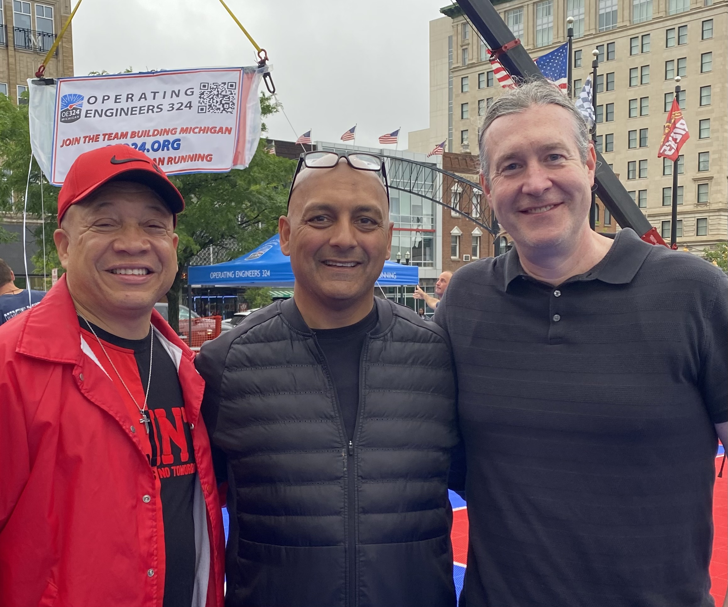 Dayne is stood to the right of two men, all are smiling into the camera. Behind there is a sign strung up between buildings which reads 'Join the Team Building Michigan'