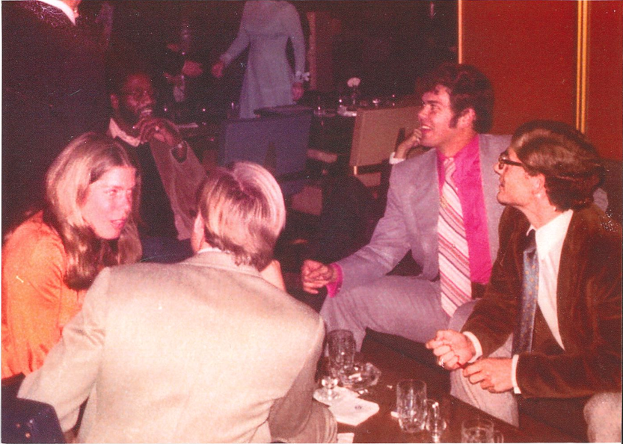 A group of people sitting around a table. There are glasses on the table and they all seem to be talking to each other. Their attire is formal.